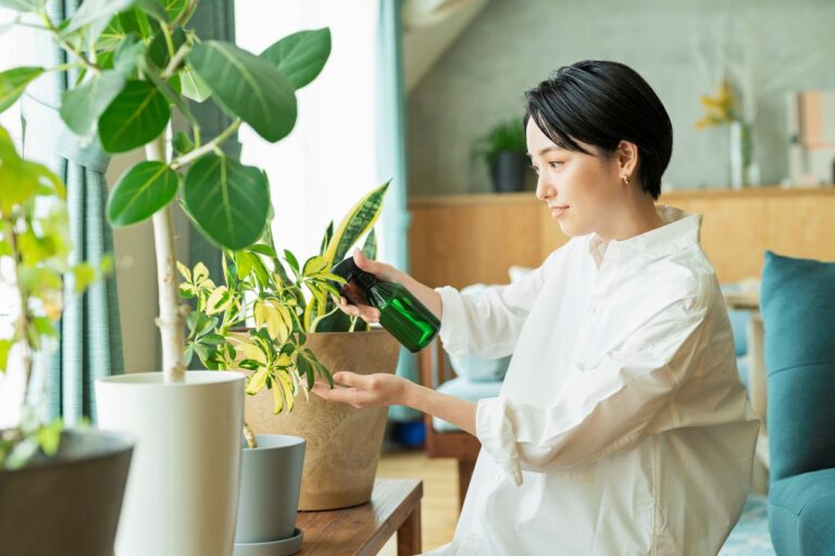 A young woman in a white shirt uses a green spray bottle to mist houseplants in a bright apartment.