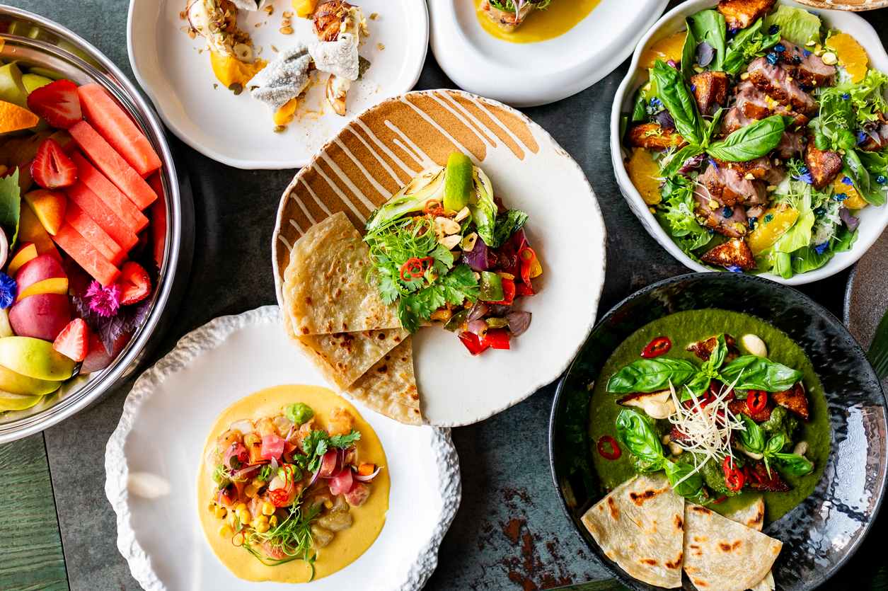 Seven food dishes with various foods, including salads, fruit, and pita.