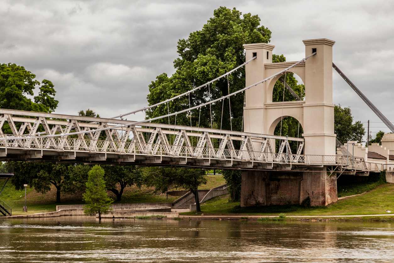 Waco suspension bridge crossing the Brazos River with trees and green lawns in the background and a cloudy sky.
