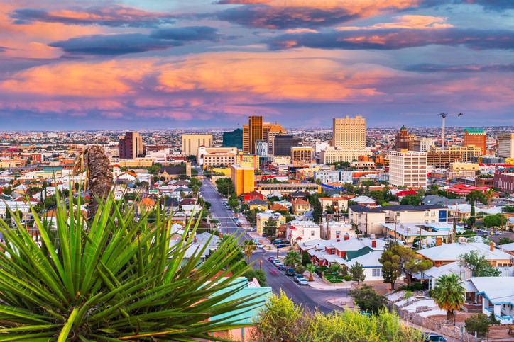 Downtown El Paso skyline at sunset with illuminated high-rise buildings, residential homes in the foreground, and the Franklin Mountains in the background.